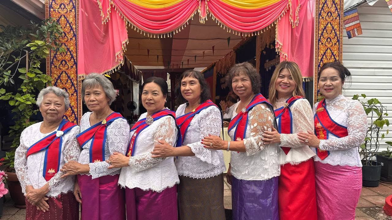 A line of women in white lace blouses, red and blue sashes and silk skirts, smiling in front of a pink and gold canopy