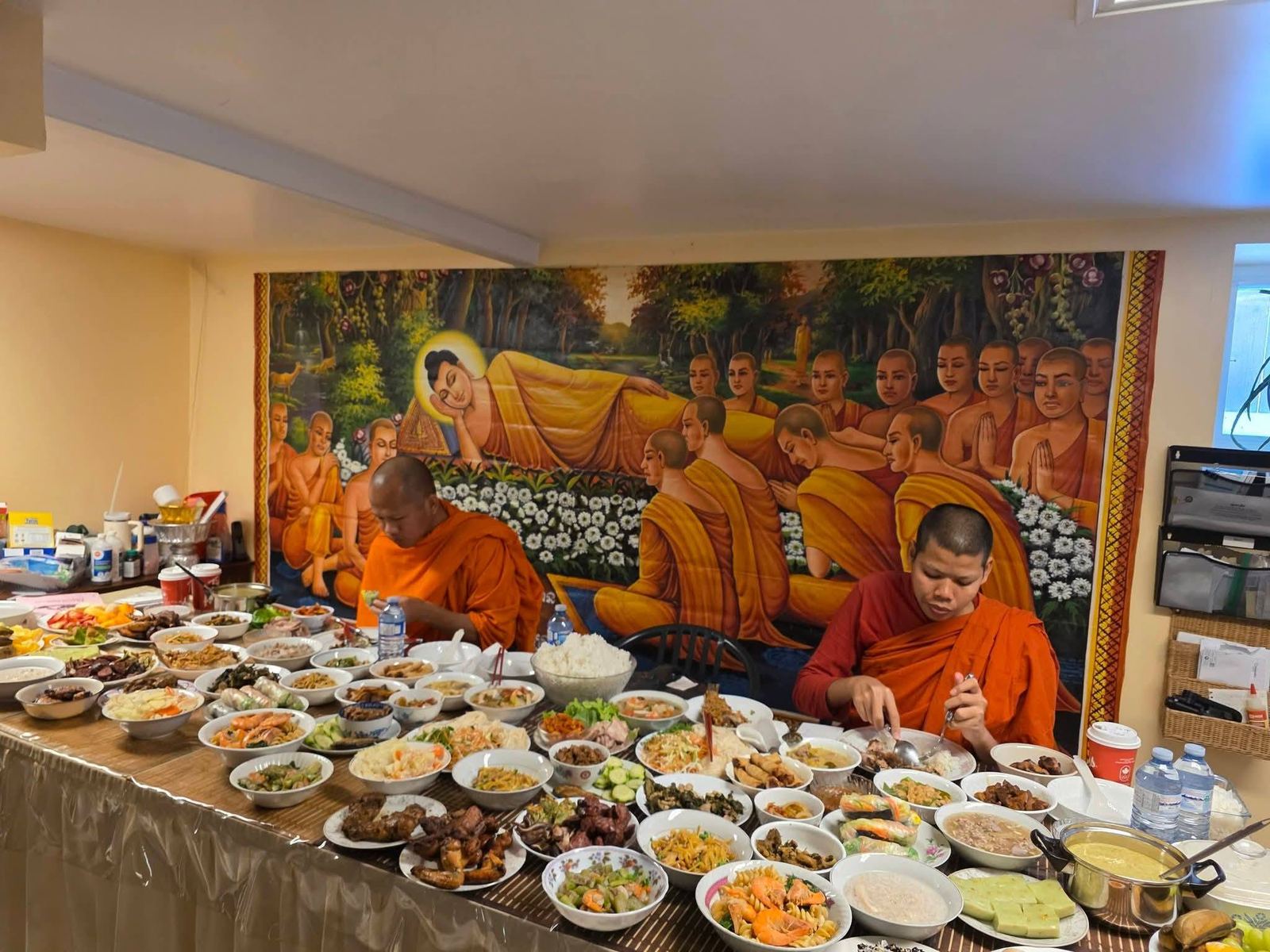 Two monks seated at a long table of home-cooked dishes offered by the community, in front of a mural of the reclining Buddha