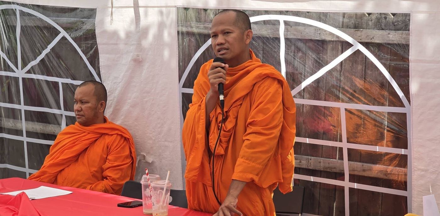 A monk in saffron robes speaking into a microphone, with a second monk seated beside him
