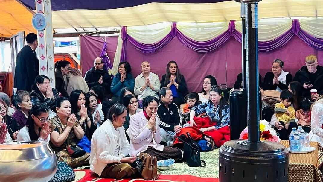 A crowd praying together under a purple and cream draped ceiling, an alms bowl in the foreground