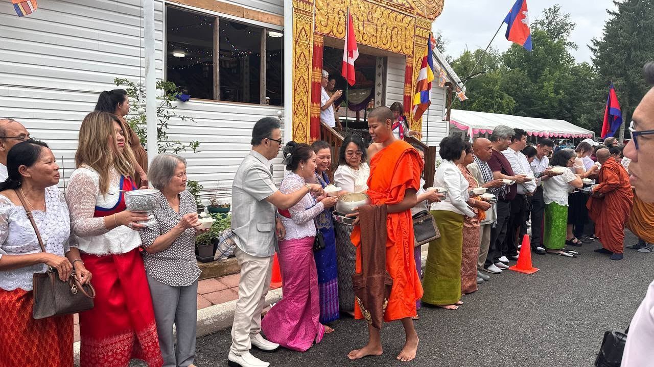 Families in traditional dress offering food to a monk in saffron robes outside the temple