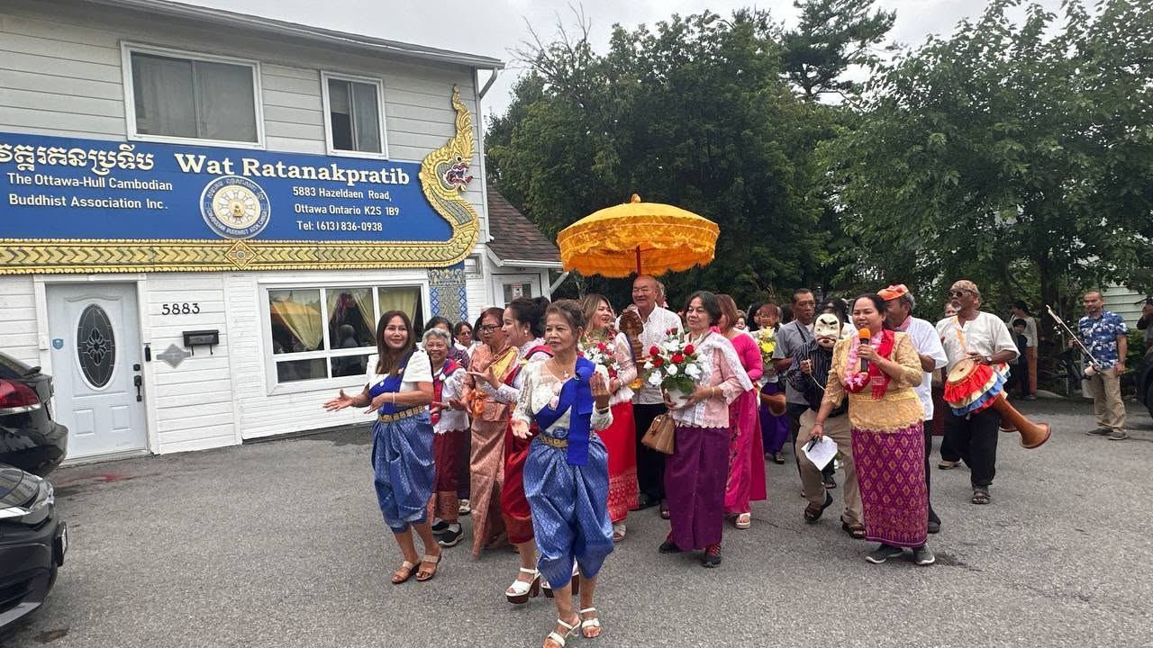 A procession in traditional Khmer dress walking under a golden umbrella in front of the temple sign